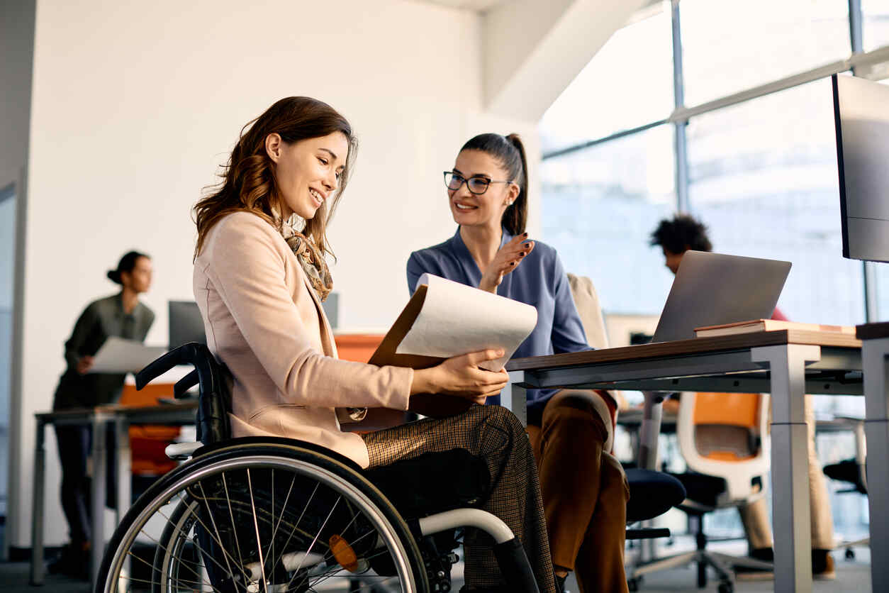 Woman in wheelchair smiling while reviewing documents with a colleague, symbolizing workplace accommodation.