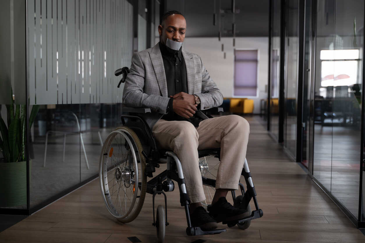 A man with a disability, gagged and hands tied while in a wheelchair in a modern office, representing the silencing of disabled employees.