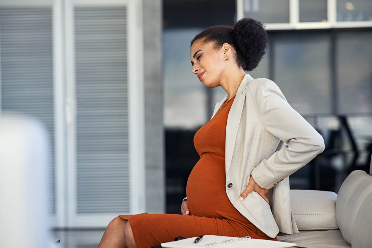 A professionally dressed pregnant woman sitting on a couch and showing signs of distress, representing the challenges of pregnancy in the workplace.